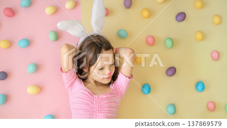 Cute child wearing bunny ears with painted face posing among colorful Easter eggs. Concept of Easter celebration, spring holiday, childhood joy and festive decorations. Cute child wearing bunny ears with painted face posing among colorful Easter eggs. Concept of Easter celebration, spring holiday, childhood joy and festive decorations. 137869579
