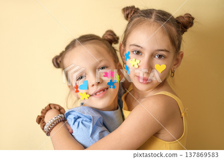 Kind friendly girls with a face decorated with pieces of colored puzzles. Children with autism hug each other, showing their love and care. Kind friendly girls with a face decorated with pieces of colored puzzles. Children with autism hug each other, showing their love and care. 137869583