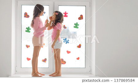 The girls are standing on the windowsill, jointly decorating the window glass with puzzle pieces and hearts.  A banner in support of people with autism spectrum disorder. 137869584
