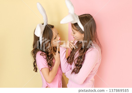 Two girls wearing bunny ears and Easter face paint smiling and looking at each other on pastel background. Concept of friendship, childhood, spring holiday and Easter celebration. 137869585