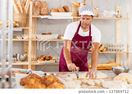 Caucasian man work in bakery as baker, cut dough into portions, forms pieces of dough to create buns Caucasian man work in bakery as baker, cut dough into portions, forms pieces of dough to create buns 137869712