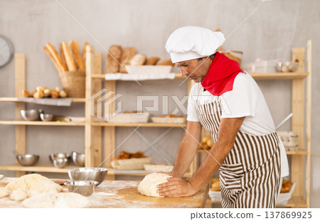 Male baker stands at work bench, kneading and shaping dough to make bread, croissants and baguettes Male baker stands at work bench, kneading and shaping dough to make bread, croissants and baguettes 137869925