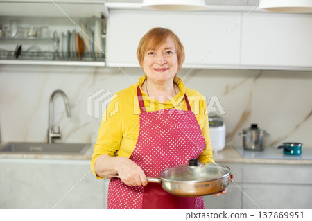Elderly woman in polka dot apron stands near kitchen table and holds frying pan 137869951