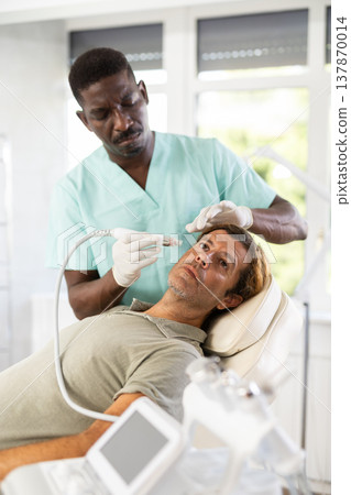 African male cosmetologist performs skin cleansing procedure for man client with beauty machine 137870014