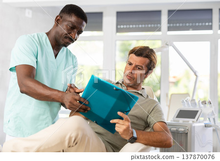 African doctor shows client papers for signing before rejuvenating procedure using beauty machine. African doctor shows client papers for signing before rejuvenating procedure using beauty machine. 137870076