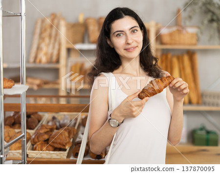 Young female customer stands with croissant in hands near window of bakery. Young female customer stands with croissant in hands near window of bakery. 137870095