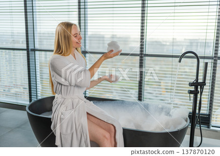 Spa relaxation. Young woman in spa bathrobe sitting at bath with foam in bathtub. Girl relaxing in bathroom at home. Female spending morning in bathroom. Beauty treatment body skin care concept Spa relaxation. Young woman in spa bathrobe sitting at bath with foam in bathtub. Girl relaxing in bathroom at home. Female spending morning in bathroom. Beauty treatment body skin care concept 137870120