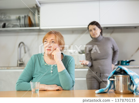 Sad mature woman with soup in pan on table listens unpleasant sayings of adult daughter. 137870135