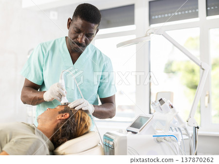 African male specialist uses ultrasonic import nozzle to care man client face skin African male specialist uses ultrasonic import nozzle to care man client face skin 137870262