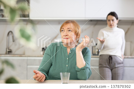 Elderly sad woman sits at table in kitchen and listens to insults.Concept stream of abuse 137870302