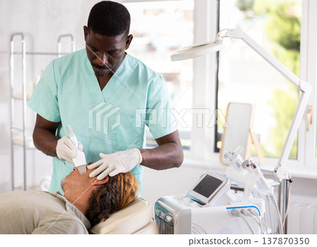 African male doctor uses scriber beauty machine nozzle for gentle skin peeling for man client African male doctor uses scriber beauty machine nozzle for gentle skin peeling for man client 137870350