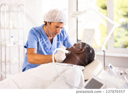 Male cosmetologist performs skin cleansing procedure for African man client with beauty machine 137870357