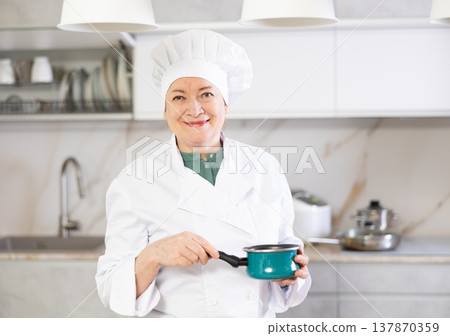Happy mature female cook in uniform standing and showing her kitchen 137870359