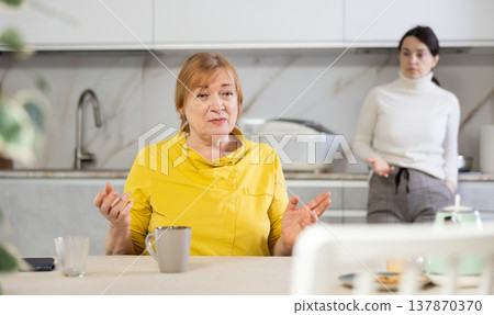 Elderly sad woman sits at table in kitchen and listens to insults.Concept stream of abuse Elderly sad woman sits at table in kitchen and listens to insults.Concept stream of abuse 137870370