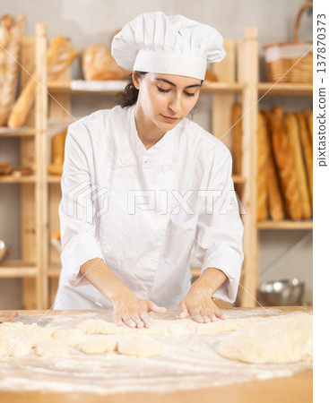 Young woman works as baker, mold baguette from dough, creates long loafs from dough. Young woman works as baker, mold baguette from dough, creates long loafs from dough. 137870373