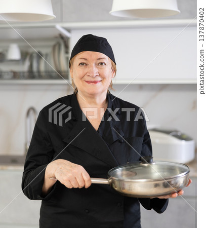 Confident mature woman in cook outfit showing around hew kitchen Confident mature woman in cook outfit showing around hew kitchen 137870402