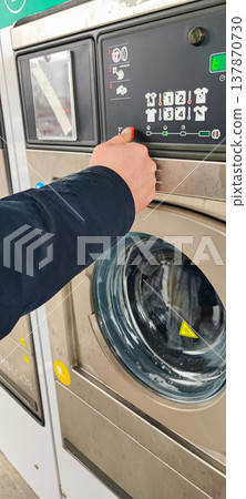 A man presses a button on the control panel of a washing machine at a self-service laundromat. Concepts of everyday life, automation, household services, and the use of modern household appliances A man presses a button on the control panel of a washing machine at a self-service laundromat. Concepts of everyday life, automation, household services, and the use of modern household appliances 137870730