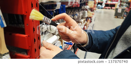 A customer holds a paint tray and brushes while shopping in a hardware store aisle. Concepts of home improvement, DIY renovation, consumer choice, and retail shopping behavior. 137870740