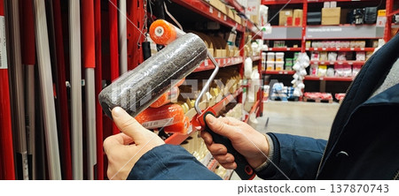 A customer holds a paint tray and brushes while shopping in a hardware store aisle. Concepts of home improvement, DIY renovation, consumer choice, and retail shopping behavior. A customer holds a paint tray and brushes while shopping in a hardware store aisle. Concepts of home improvement, DIY renovation, consumer choice, and retail shopping behavior. 137870743