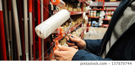 A customer holds a paint tray and brushes while shopping in a hardware store aisle. Concepts of home improvement, DIY renovation, consumer choice, and retail shopping behavior. A customer holds a paint tray and brushes while shopping in a hardware store aisle. Concepts of home improvement, DIY renovation, consumer choice, and retail shopping behavior. 137870744
