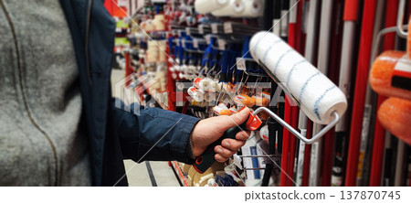 A customer holds a paint tray and brushes while shopping in a hardware store aisle. Concepts of home improvement, DIY renovation, consumer choice, and retail shopping behavior. 137870745