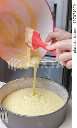 Person pouring cake batter from a mixing bowl into a round springform pan on a kitchen countertop with appliances in the background 137871029