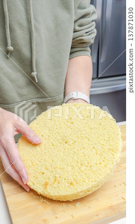 Person holding a freshly baked round sponge cake on a wooden cutting board in a modern kitchen setting with stainless steel appliances in the background Person holding a freshly baked round sponge cake on a wooden cutting board in a modern kitchen setting with stainless steel appliances in the background 137871030