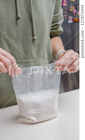 Person in green hoodie holds a transparent bag filled with white powder on a kitchen countertop with a refrigerator in the background Person in green hoodie holds a transparent bag filled with white powder on a kitchen countertop with a refrigerator in the background 137871045