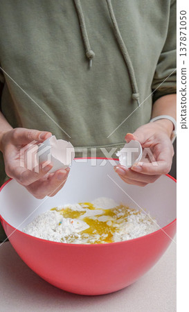 Person in green hoodie cracks eggs into a red mixing bowl filled with flour and liquid ingredients on a kitchen countertop 137871050