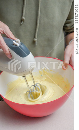 Person using electric hand mixer to blend batter in a red mixing bowl on a kitchen countertop with a neutral background Person using electric hand mixer to blend batter in a red mixing bowl on a kitchen countertop with a neutral background 137871051