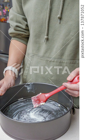 Person in green hoodie applies butter with a red brush to a round cake pan on a kitchen countertop with appliances in the background 137871052