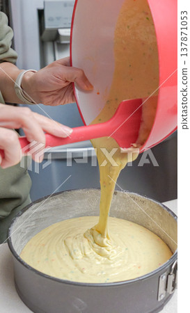 Woman pouring cake batter from a red mixing bowl into a round baking pan on a kitchen countertop with a modern appliance in the background Woman pouring cake batter from a red mixing bowl into a round baking pan on a kitchen countertop with a modern appliance in the background 137871053