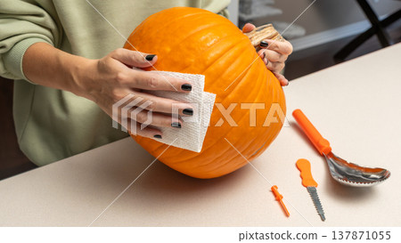Person cleaning a bright orange pumpkin with a paper towel, surrounded by carving tools on a light countertop in a cozy indoor setting Person cleaning a bright orange pumpkin with a paper towel, surrounded by carving tools on a light countertop in a cozy indoor setting 137871055