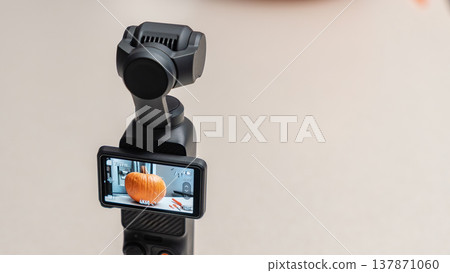 Camera stabilizer with smartphone display showing a pumpkin being cut for Halloween preparation on a clean, light-colored surface 137871060
