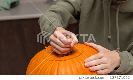 Hands of a person in a green hoodie carving the top of a bright orange pumpkin for Halloween preparation in a kitchen setting Hands of a person in a green hoodie carving the top of a bright orange pumpkin for Halloween preparation in a kitchen setting 137871062