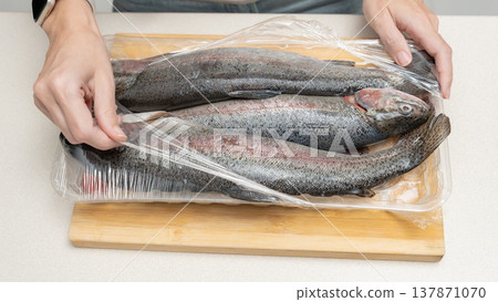 Hands removing plastic wrap from fresh rainbow trout fish on a wooden cutting board in a kitchen setting 137871070
