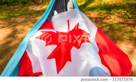 Canadian flag draped over a person relaxing in a hammock in a natural outdoor setting, surrounded by greenery and sunlight filtering through trees Canadian flag draped over a person relaxing in a hammock in a natural outdoor setting, surrounded by greenery and sunlight filtering through trees 137871076