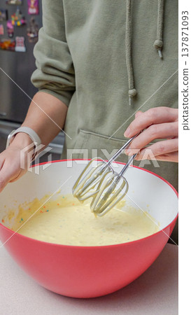 Person mixing batter in a large red bowl with electric beaters, kitchen appliances and colorful magnets visible in the background Person mixing batter in a large red bowl with electric beaters, kitchen appliances and colorful magnets visible in the background 137871093