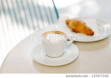 Cup of cappuccino and a croissant on a table in a street cafe. Selective focus. Cup of cappuccino and a croissant on a table in a street cafe. Selective focus. 137871549