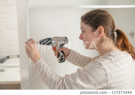 A Caucasian woman uses a screwdriver to tighten a screw while assembling a cabinet. A Caucasian woman uses a screwdriver to tighten a screw while assembling a cabinet. 137873449