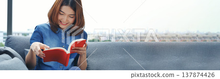Banner Relax asian woman reading book sitting on sofa in living room holding book. Banner Young woman reading open book leisure mind. Happiness beauty person smiling face happy time with copy space 137874426