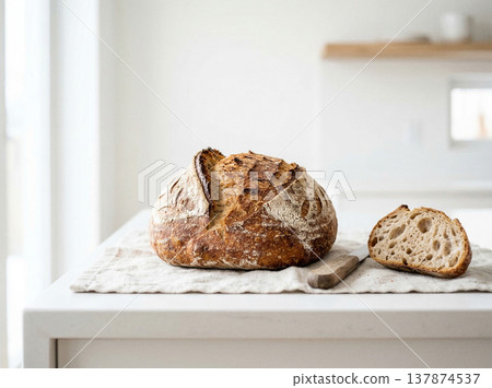Rustic sourdough bread loaf and slice resting on a linen napkin in a bright kitchen setting with baked goods Rustic sourdough bread loaf and slice resting on a linen napkin in a bright kitchen setting with baked goods 137874537