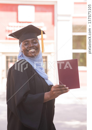 African woman in hijab and graduation gown holding diploma.  137875105