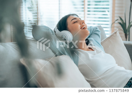 Portrait of Asian woman relaxing listening audio with headphones sitting on a couch at home Portrait of Asian woman relaxing listening audio with headphones sitting on a couch at home 137875849