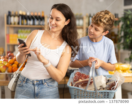 Adult woman with phone and son choosing groceries in store Adult woman with phone and son choosing groceries in store 137878090
