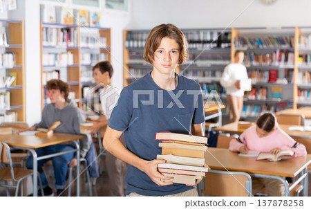 Teenager boy standing with a stack of books in library 137878258