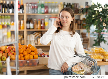 In shop teenage girl with basket in hands talks on phone, calls mother In shop teenage girl with basket in hands talks on phone, calls mother 137878395