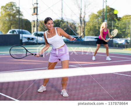Beautiful young woman hitting ball with racket, tennis players play in court Beautiful young woman hitting ball with racket, tennis players play in court 137878664