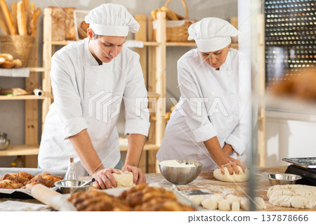 Two bakers, woman and young guy, knead raw dough on table in bakery kitchen 137878666