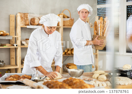 Female baker kneads dough on kitchen counter while young male assistant carries fresh baguettes to bakery display case Female baker kneads dough on kitchen counter while young male assistant carries fresh baguettes to bakery display case 137878761